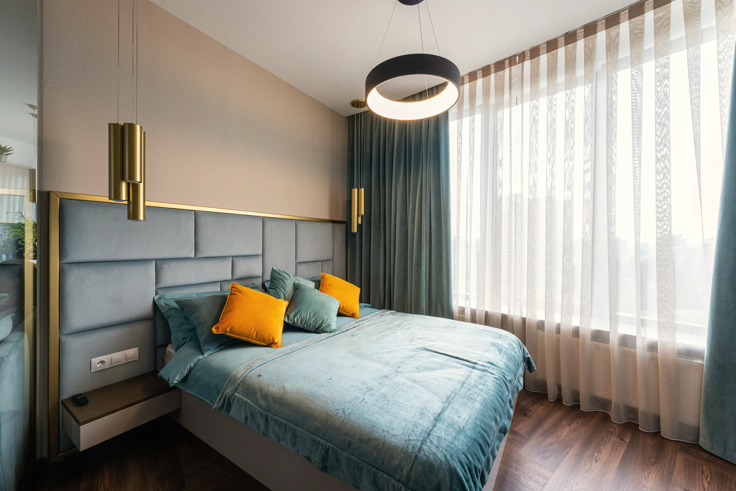Example bedroom: an upholstered headboard wall framed in brass, brass bedside pendants and layered sheer and velvet curtains.